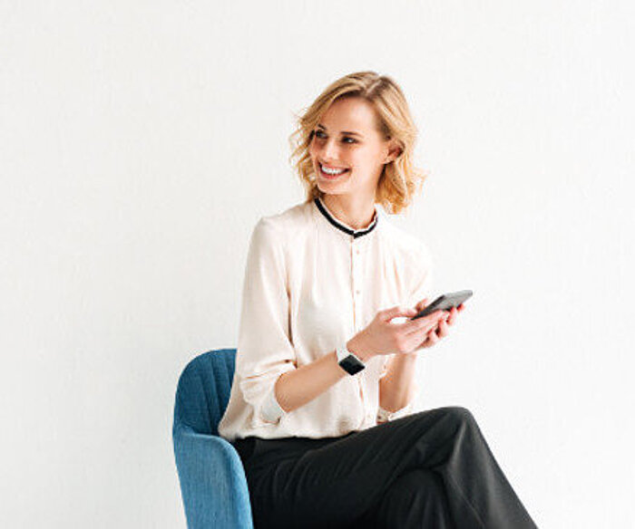 Smiling blonde woman in cream blouse sitting on blue chair, holding smartphone, wearing smartwatch