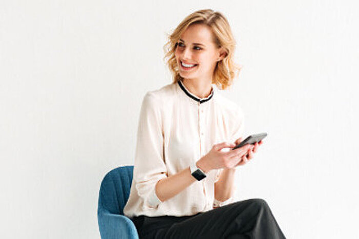 Smiling blonde woman in cream blouse sitting on blue chair, holding smartphone, wearing smartwatch