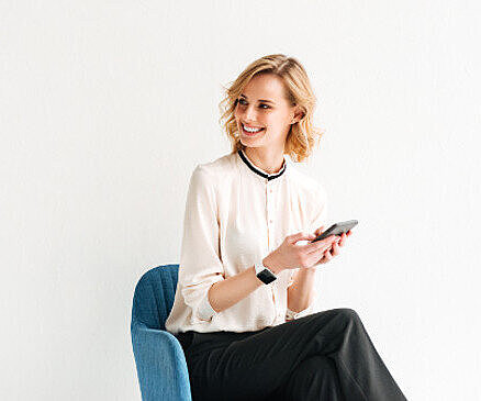 Smiling blonde woman in cream blouse sitting on blue chair, holding smartphone, wearing smartwatch