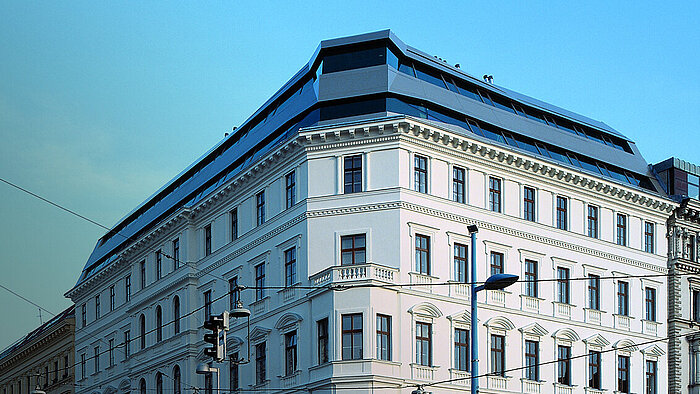 Klassizistisches weißes Eckgebäude mit modernem verglastem Dachaufbau vor blauem Himmel, Straßenlaterne und Oberleitungen