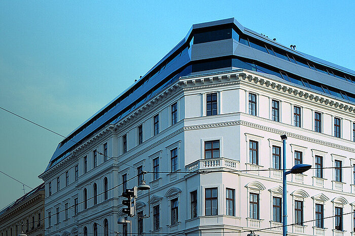 Klassizistisches weißes Eckgebäude mit modernem verglastem Dachaufbau vor blauem Himmel, Straßenlaterne und Oberleitungen