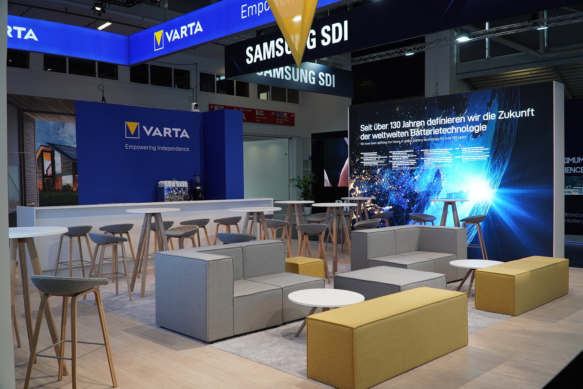 Trade fair lounge with grey sofas, yellow benches and bar stools at VARTA and Samsung SDI battery technology booths
