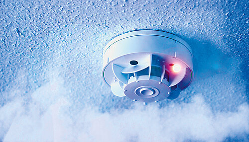 Ceiling-mounted smoke detector with red indicator light, surrounded by smoke against a blue-tinted background