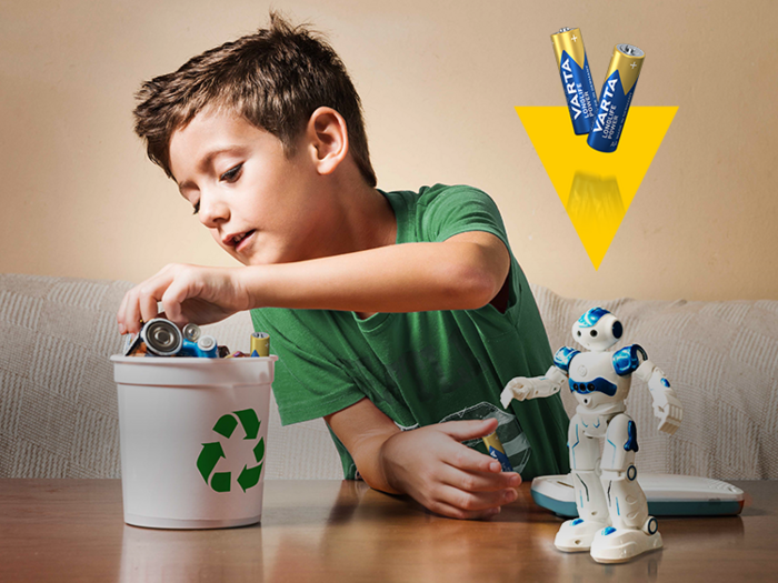 Junge wirft Batterien in einen Recyclingeimer, daneben ein Spielzeugroboter und Varta-Batterien