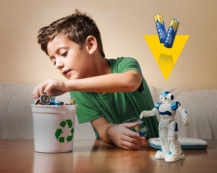 Boy placing batteries into a recycling bin next to a white toy robot, with Varta batteries shown above
