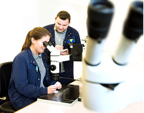 Two people in dark blue lab coats work at a microscope; woman looks through eyepiece while man takes notes beside her