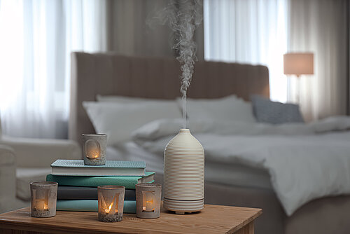 White ceramic aroma diffuser emitting steam beside lit candles and stacked books on a wooden table in a bedroom