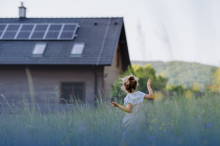 Mädchen in hellem Kleid läuft durch hohe Wiese vor Holzhaus mit Solaranlage auf dem Dach