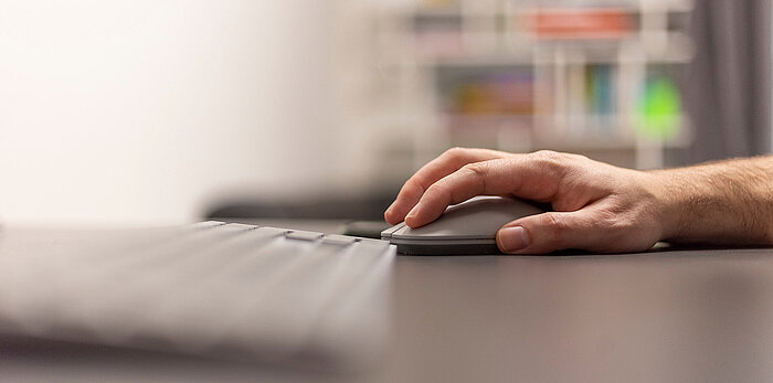 Close-up of a man's hand resting on a gray wireless mouse beside a keyboard on a dark desk