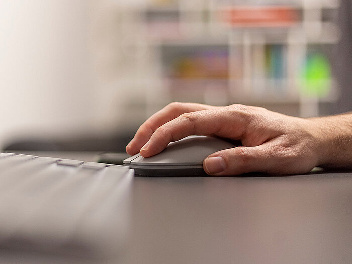 Close-up of a man's hand resting on a gray wireless mouse beside a keyboard on a dark desk