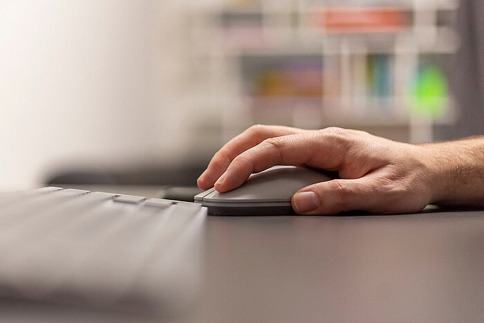 Nærbilde av en hånd som hviler på en grå datamus ved siden av et tastatur på et mørkt skrivebord