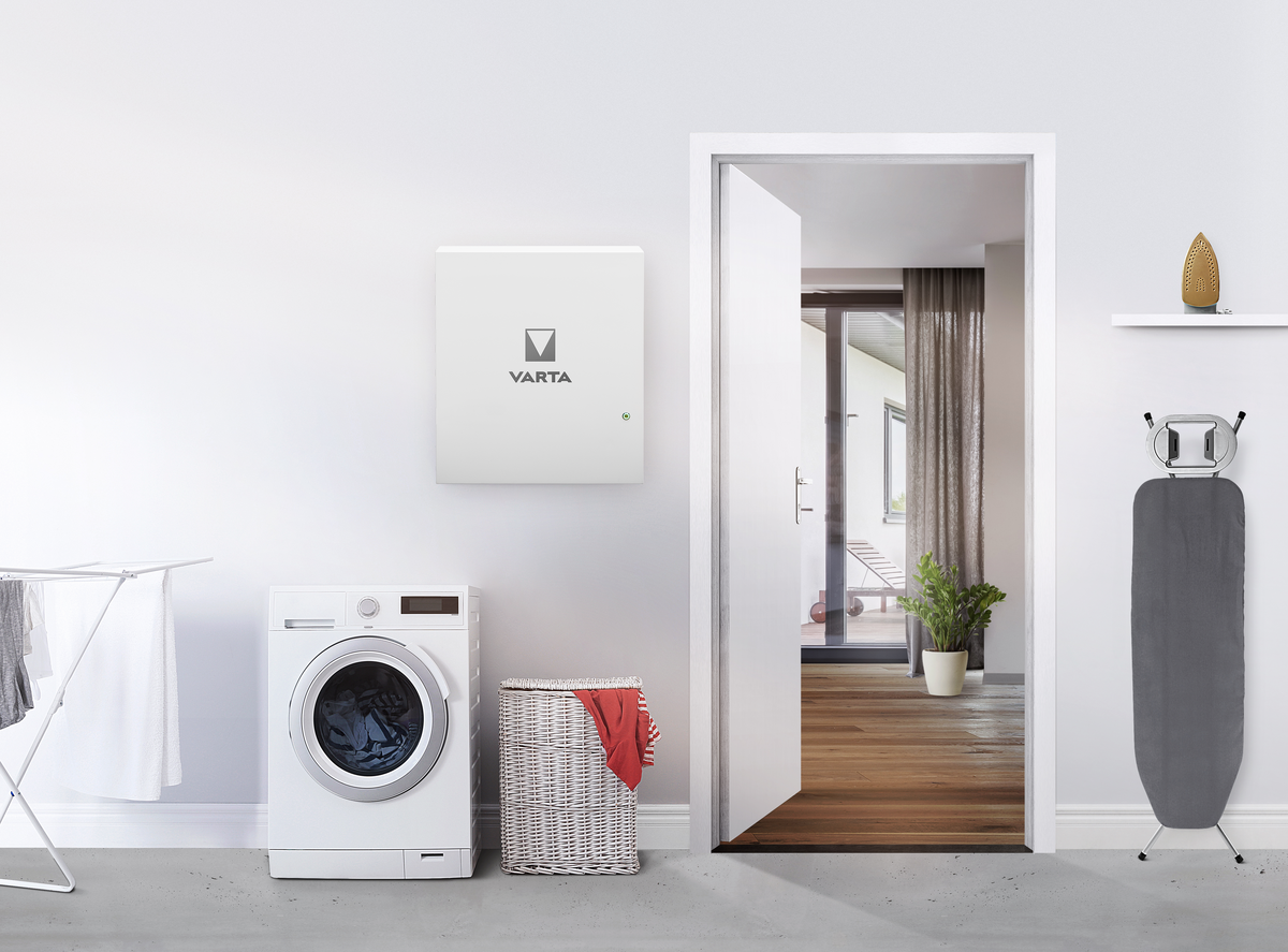 Wall-mounted white VARTA energy storage unit in a laundry room with washing machine, basket, drying rack and ironing board