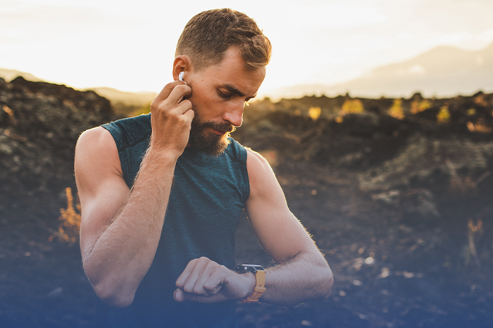 Bärtiger Mann mit Sportshirt setzt Earbuds ein und schaut auf seine Smartwatch in hügeliger Landschaft bei Sonnenuntergang