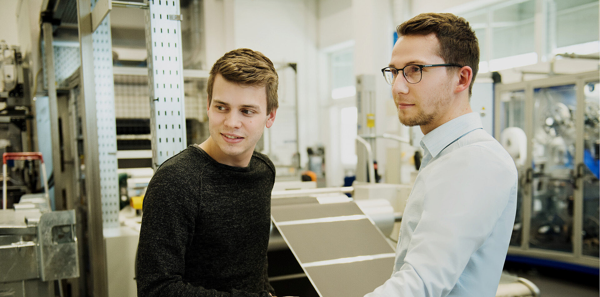 Two young men converse in an industrial facility with machinery, one in a dark sweater, the other in a light shirt and glasses