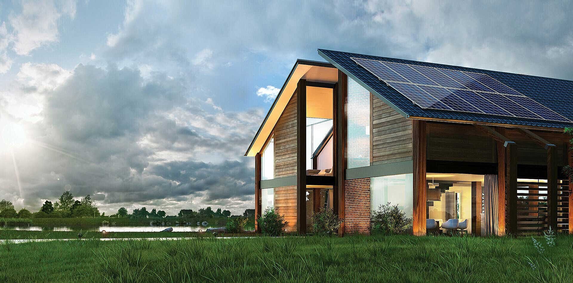 Modern wooden barn-style house with large solar panels on sloped roof, illuminated interior, beside lake under cloudy sky
