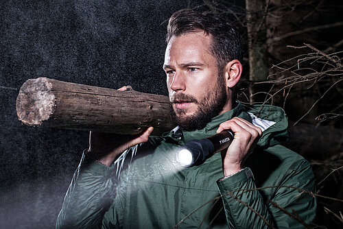 Bearded man in green jacket carries wooden log on shoulder while holding lit flashlight at night