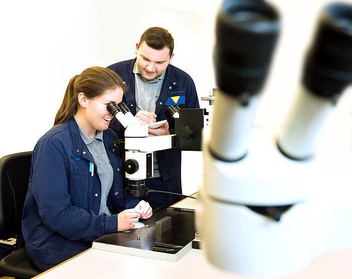 Two technicians in blue work jackets examine samples; woman looks through microscope while man takes notes beside her