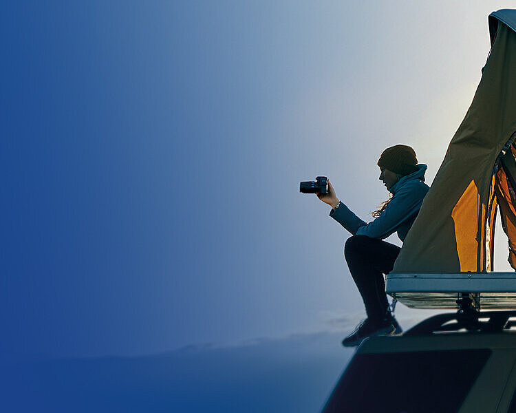 Persona con berretto seduta su tenda da tetto fotografa con fotocamera al crepuscolo contro cielo blu