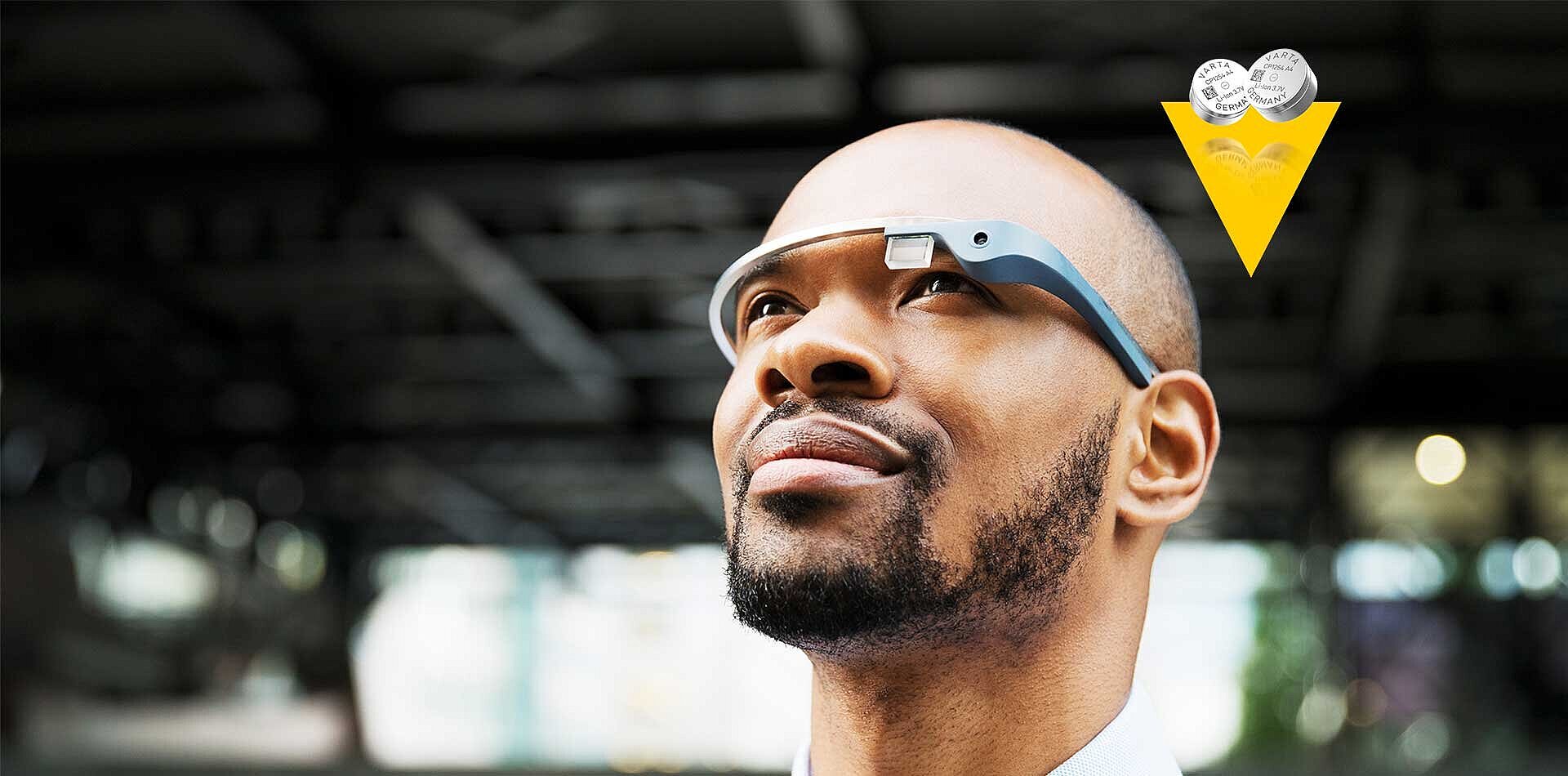 Bald man with beard wearing smart glasses looks upward, with Varta coin batteries shown in a yellow triangle graphic