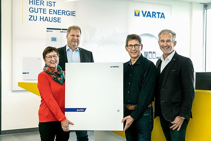 Vier lächelnde Personen halten gemeinsam einen weißen VARTA-Pulse-Batteriespeicher in einem Showroom mit Logo und Schriftzug „Hier ist gute Energie zu Hause
