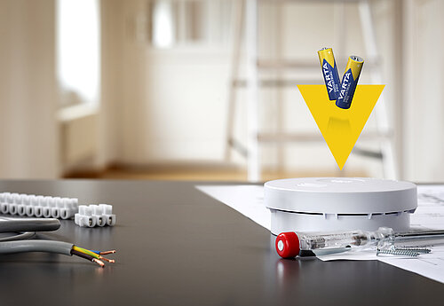 Smoke detector on table with batteries above, voltage tester and wire connectors nearby, ladder blurred in background