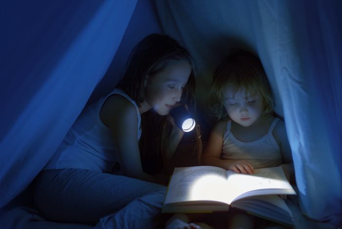 Two children read a book together under a sheet tent at night, illuminated by a flashlight held by the older girl