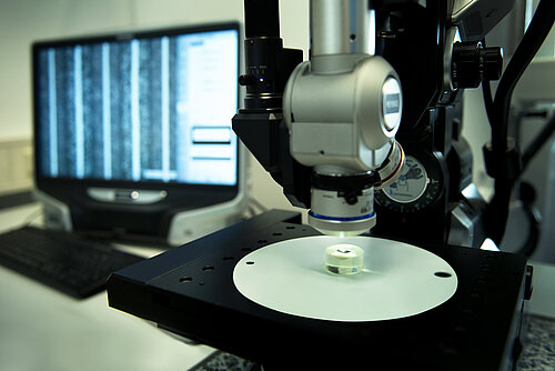 Modern laboratory microscope examining a small sample on a stage, with analysis displayed on a computer monitor behind