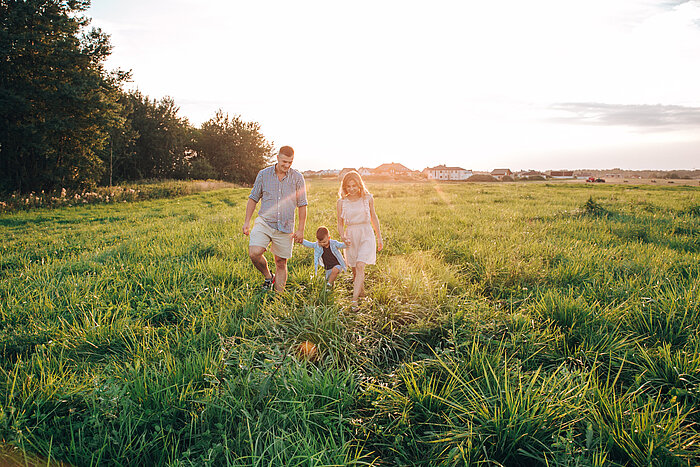 Family of three walking hand in hand through a sunlit grassy field at sunset, with houses visible in the distance