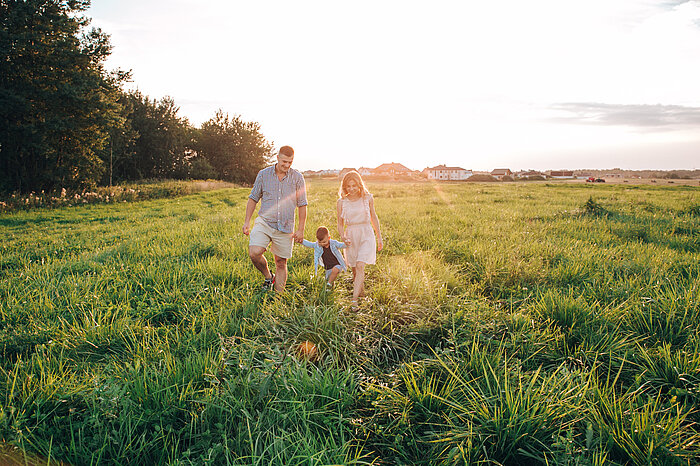 Family of three walking hand in hand through a sunlit grassy field at sunset, with houses visible in the distance