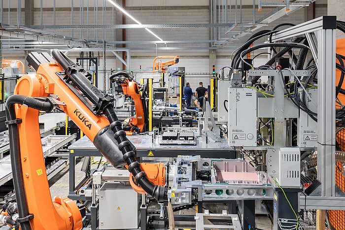 Orange KUKA industrial robot arm in a modern automated factory with assembly equipment and workers in the background