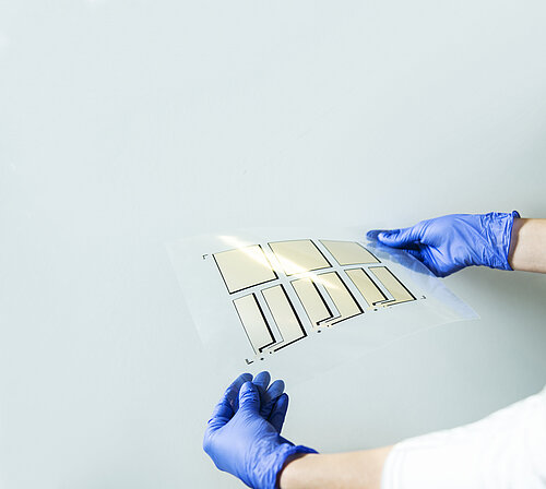Gloved hands hold a transparent flexible film with printed rectangular gold-colored electrode patterns arranged in a grid