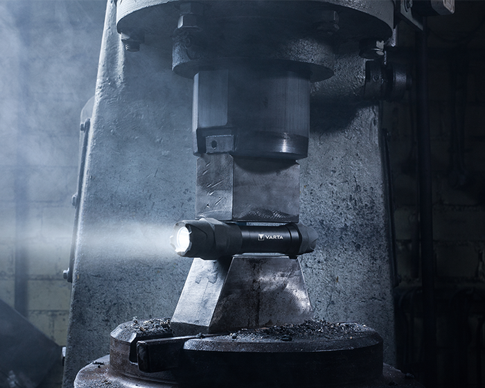 Illuminated black Varta flashlight placed between metal press components in an industrial workshop with hazy atmosphere