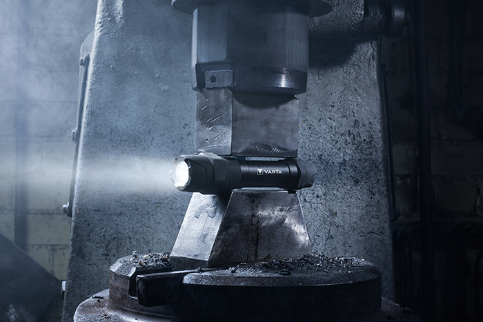 Illuminated black Varta flashlight placed between metal press components in an industrial workshop with hazy atmosphere