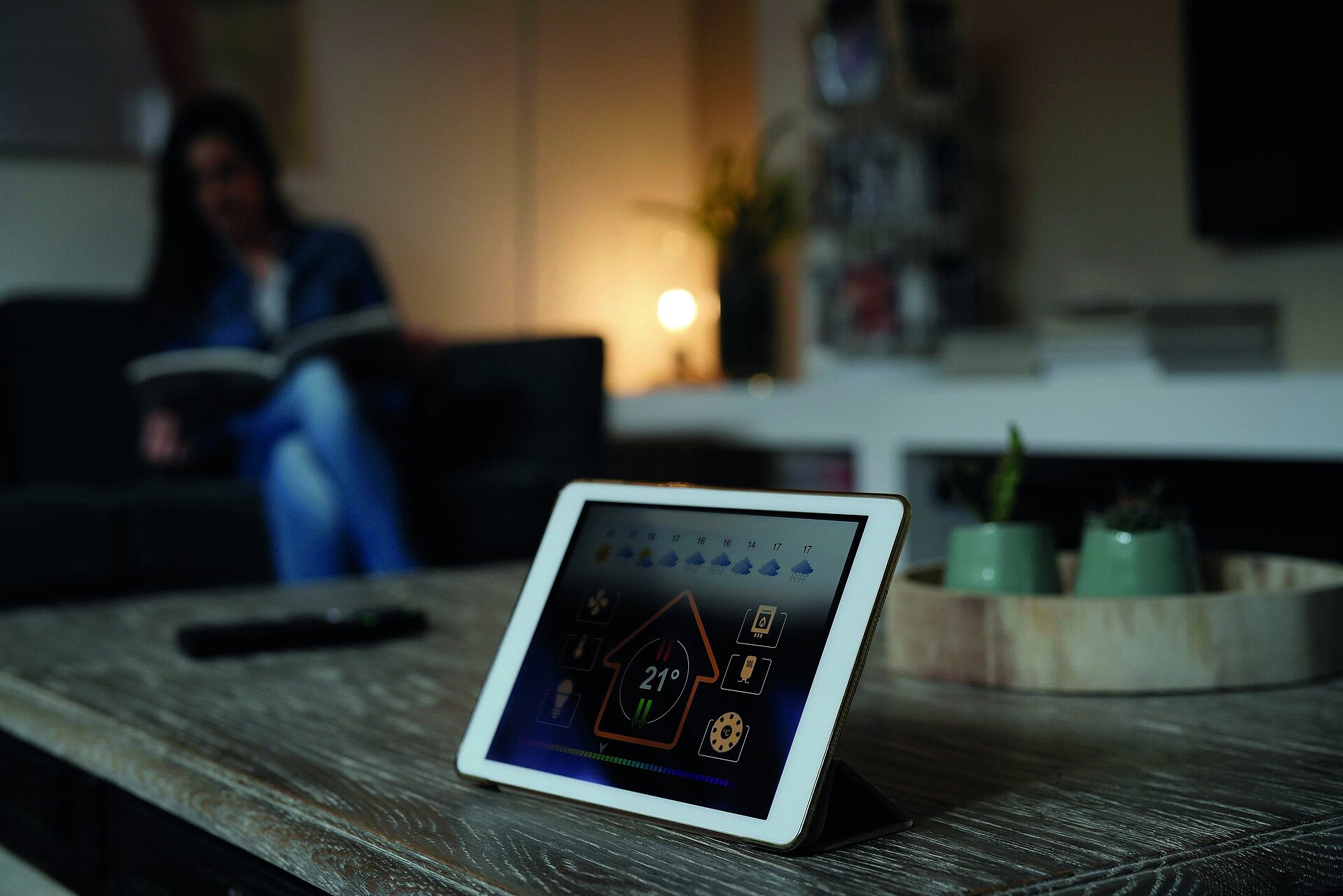 Tablet on wooden table displays smart home app at 21°C; woman reads book on sofa in blurred background