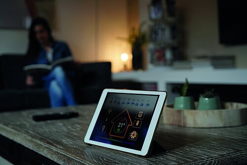 Tablet on wooden table displays smart home app at 21°C; woman reads book on sofa in blurred background