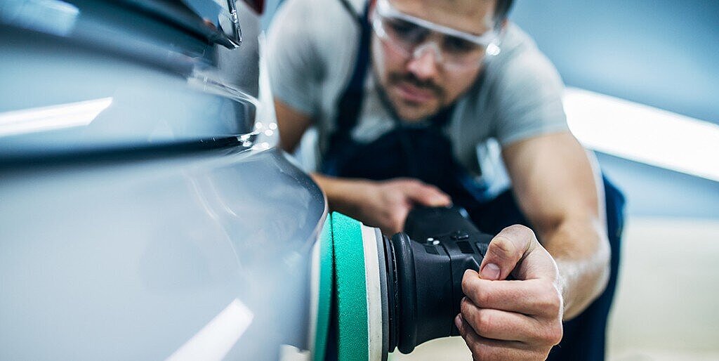 Man with safety goggles polishing a light-colored car body using a handheld dual-action polisher with green pad