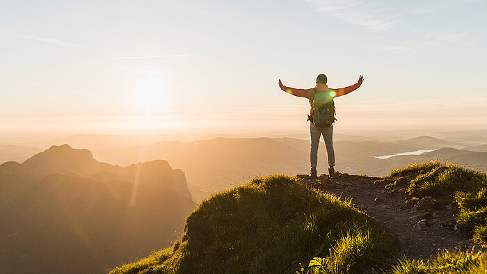 Hiker with backpack stands on mountain summit at sunrise, arms raised wide, overlooking misty valleys and distant peaks