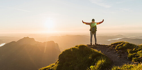 Hiker with backpack stands on mountain summit at sunrise, arms raised wide, overlooking misty valleys and distant peaks