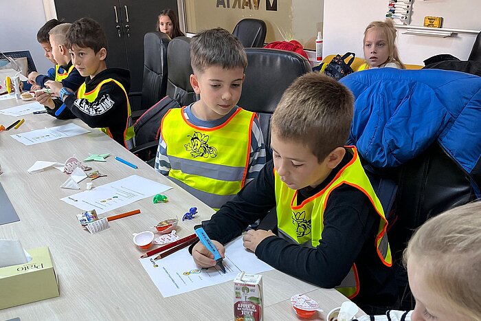 Kinder mit gelben Warnwesten sitzen an einem Tisch und malen, vor ihnen Stifte, Papiere und ein Apfelsaft-Tetrapack
