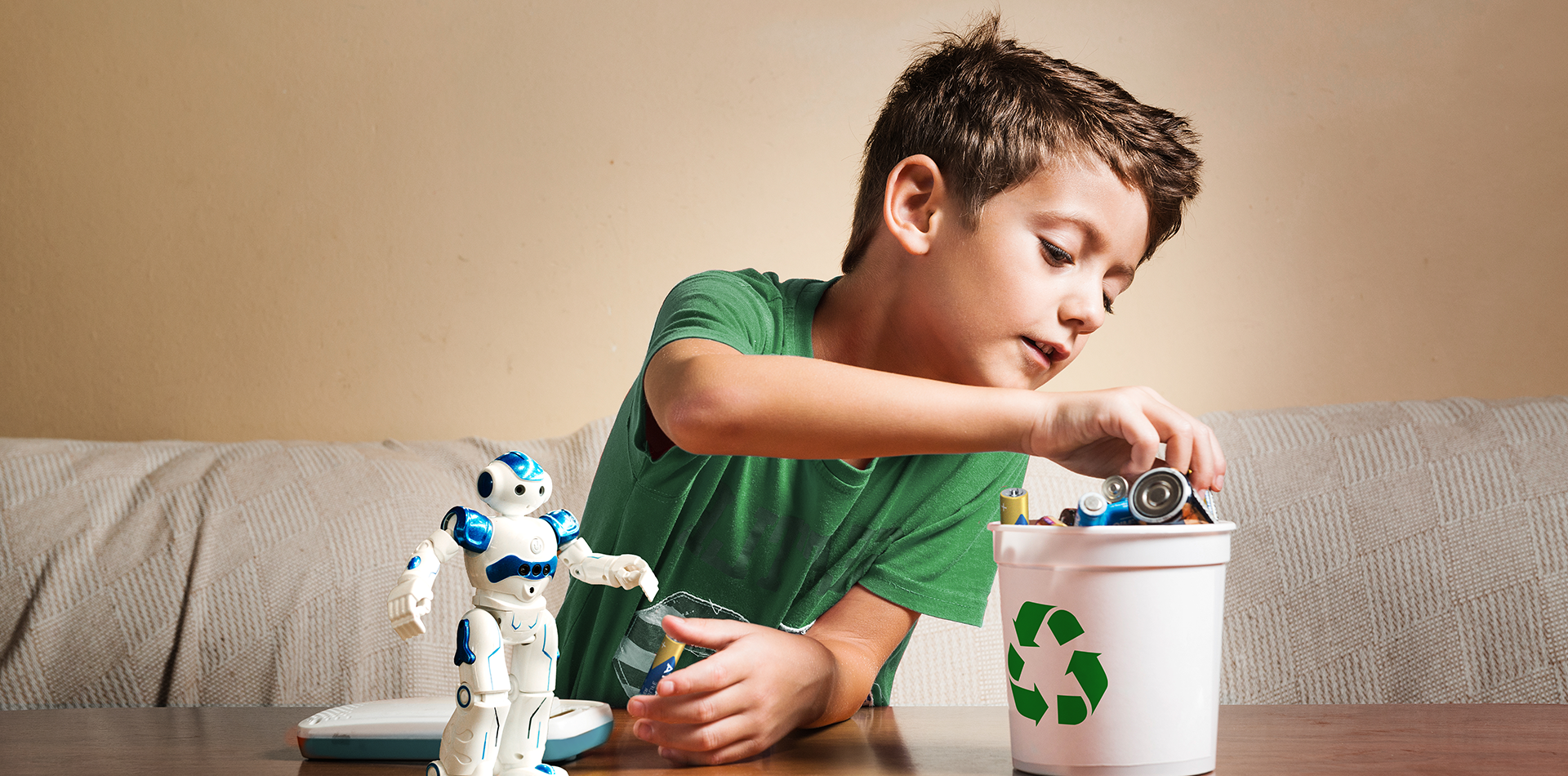 Chłopiec w zielonej koszulce wrzuca zużyte baterie do białego pojemnika z zielonym symbolem recyklingu, obok robot-zabawka