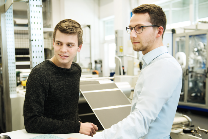 Zwei junge Männer im Gespräch in einer industriellen Produktionshalle mit Maschinen im Hintergrund