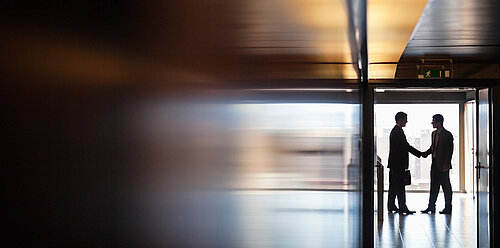 Two businessmen in suits shaking hands silhouetted against a bright window near an emergency exit sign in an office