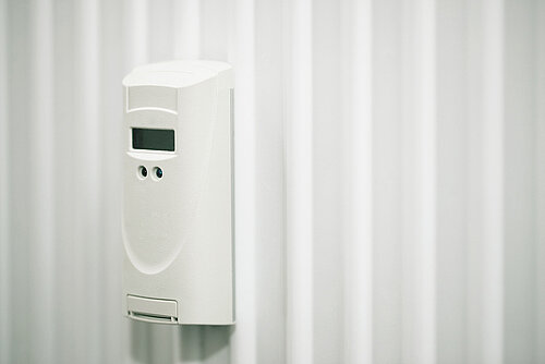 Wall-mounted white automatic air freshener dispenser with small LCD display against a light corrugated background