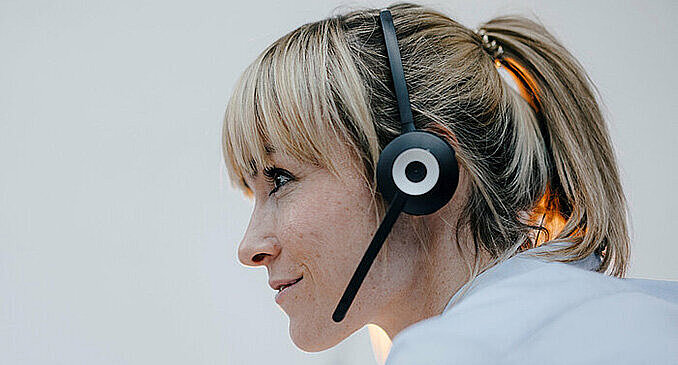 Side profile of a blonde woman with ponytail wearing a black headset, smiling slightly against a light background