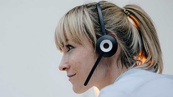 Side profile of a blonde woman with ponytail wearing a black headset, smiling slightly against a light background
