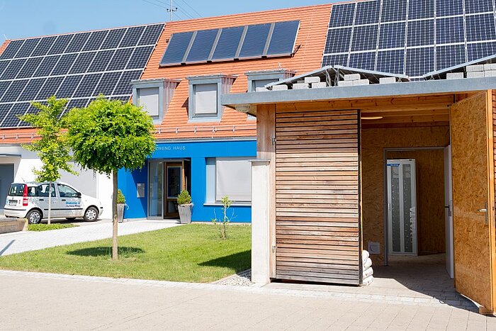 Gebäude mit Solarpaneelen auf Ziegeldach, blauer Fassade, Holzanbau und weißem Lieferwagen auf gepflastertem Vorplatz