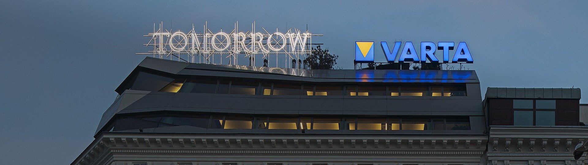 暮色中一座现代建筑屋顶上设有发光的"TOMORROW"字样霓虹灯牌和蓝色"VARTA"标志,下方楼层窗户透出灯光