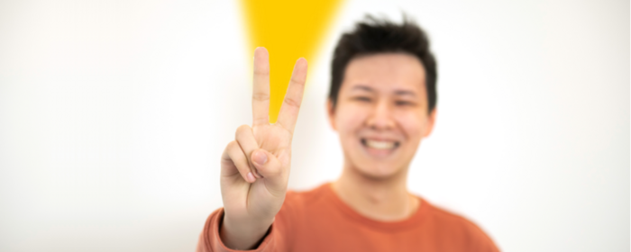 Smiling young man making a peace sign with his hand in sharp focus, wearing an orange sweater against a white background