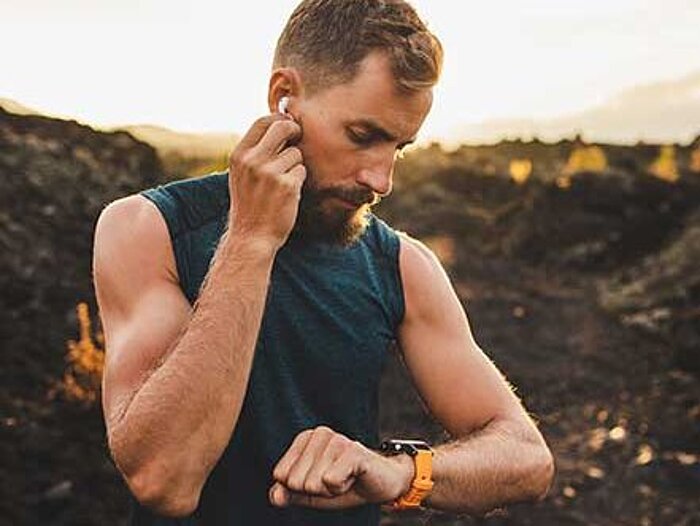 Bärtiger Mann mit ärmellosem Shirt setzt Kopfhörer ein und blickt auf Sportuhr, im Hintergrund Landschaft bei Sonnenuntergang