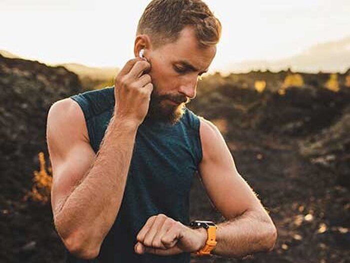 Bearded man in dark sleeveless shirt adjusts earbud while checking orange sports watch outdoors at sunset